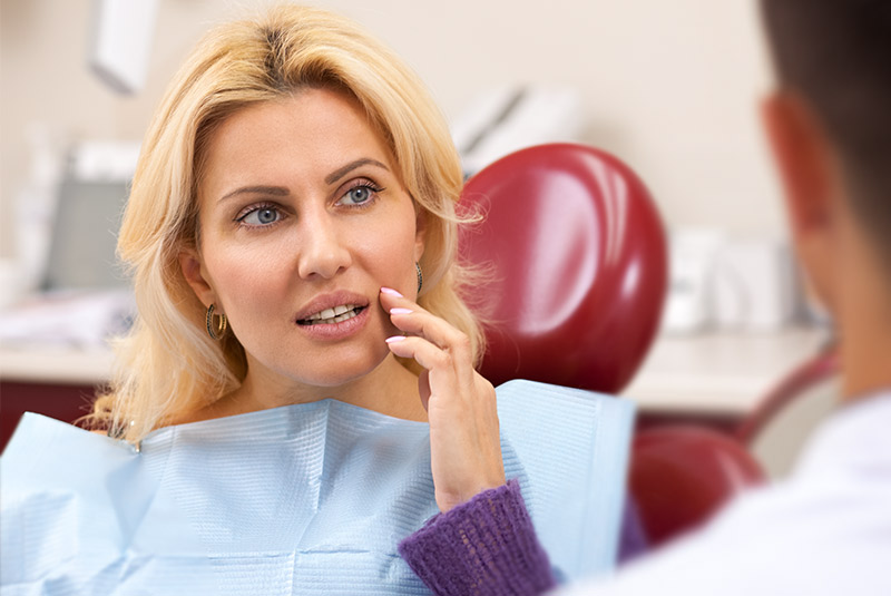 Very nice lady with blonde hair sitting in dental chair touching mouth in pain lookin at doctor