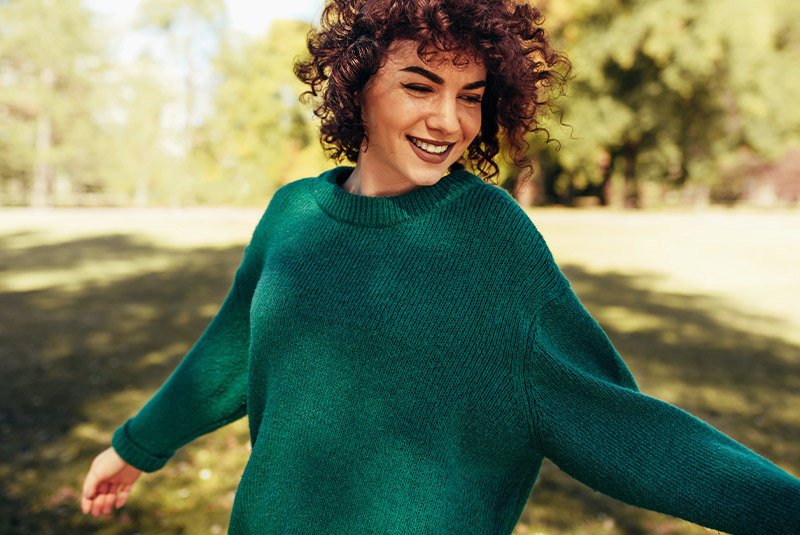 Very nice lady spinng outside grass and trees in the background she's smiling wearing green sweater with red hair